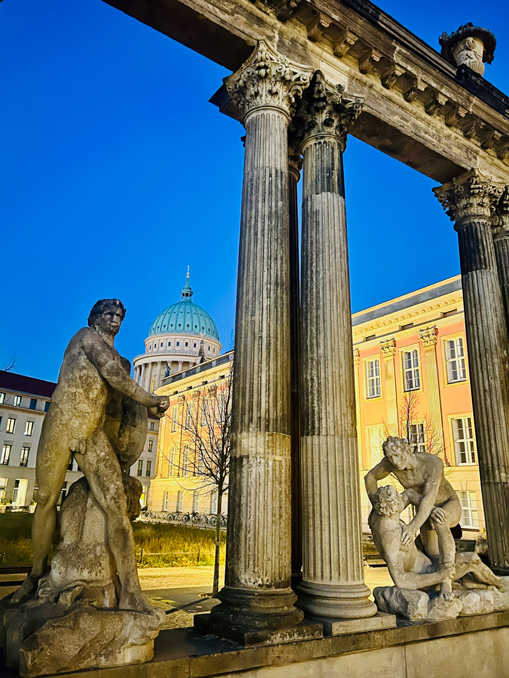 Steinfiguren unter einem Säulenportal, hinten Gebäude und eine Kuppel